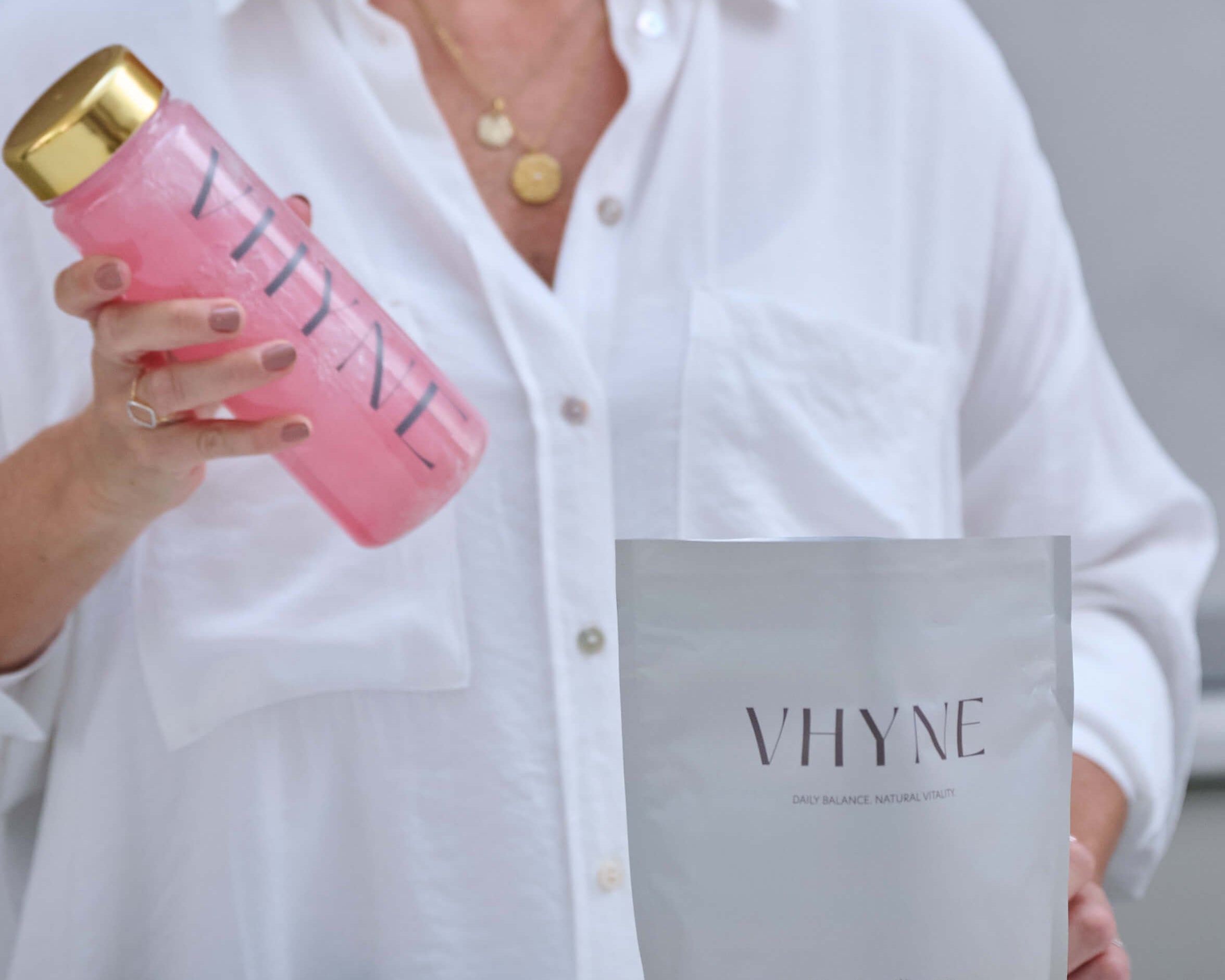 Person holding a Vhyne  branded water bottle shaking her VitaResiliance with water. An open packet of VitaResiliance is on the counter top with a gold supplement scoop in front.