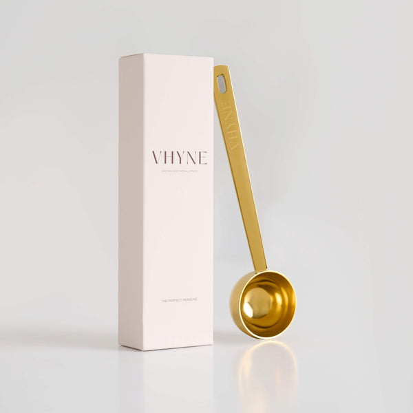 Gold measuring spoon with 'VHYNE' branding next to its packaging on a light grey background